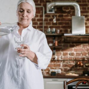 Ältere Dame giesst sich ein Glas Wasser aus Kanne in ihrer Küche ein.